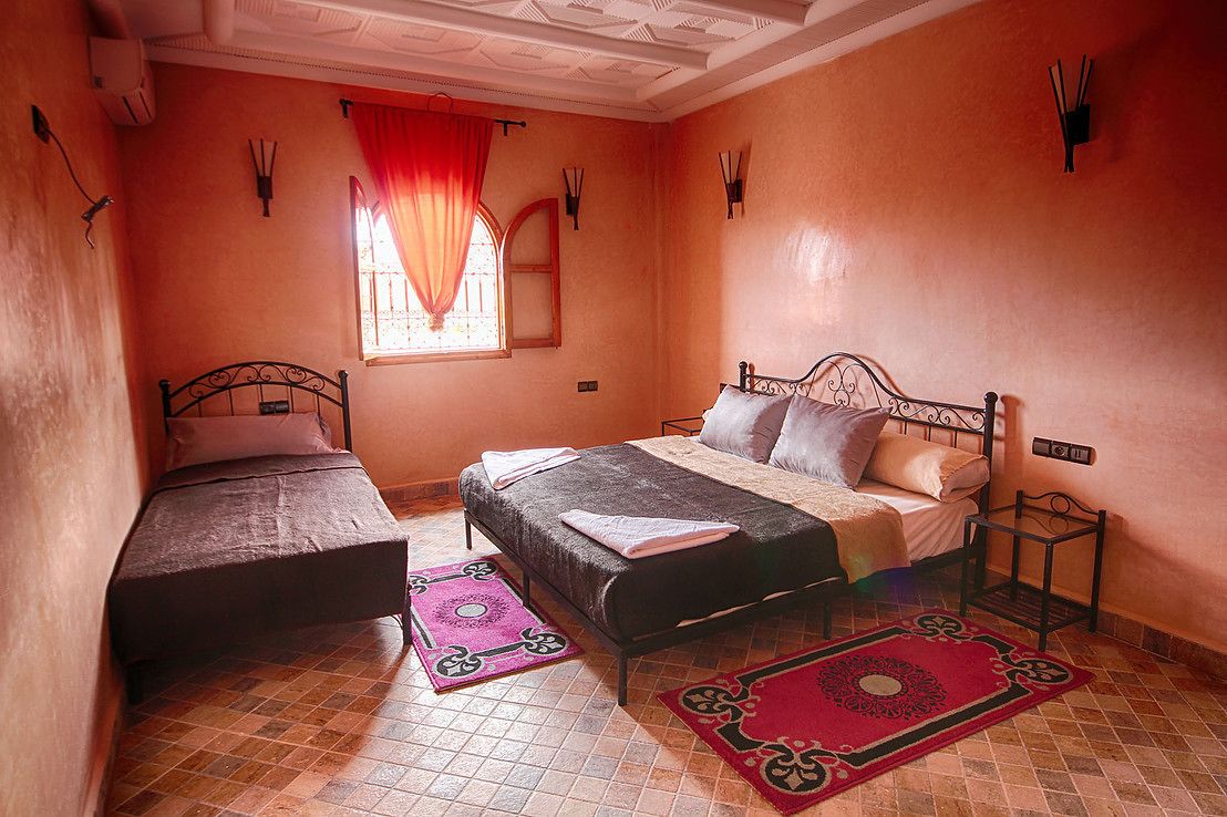 Une chambre avec deux lits, une fenêtre avec des rideaux, un sol carrelé et un luminaire fixé au mur.