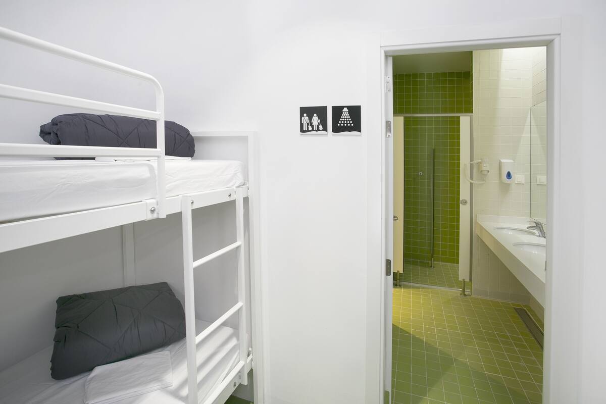 Habitación con literas y un baño con azulejos verdes visible a través de una puerta abierta.