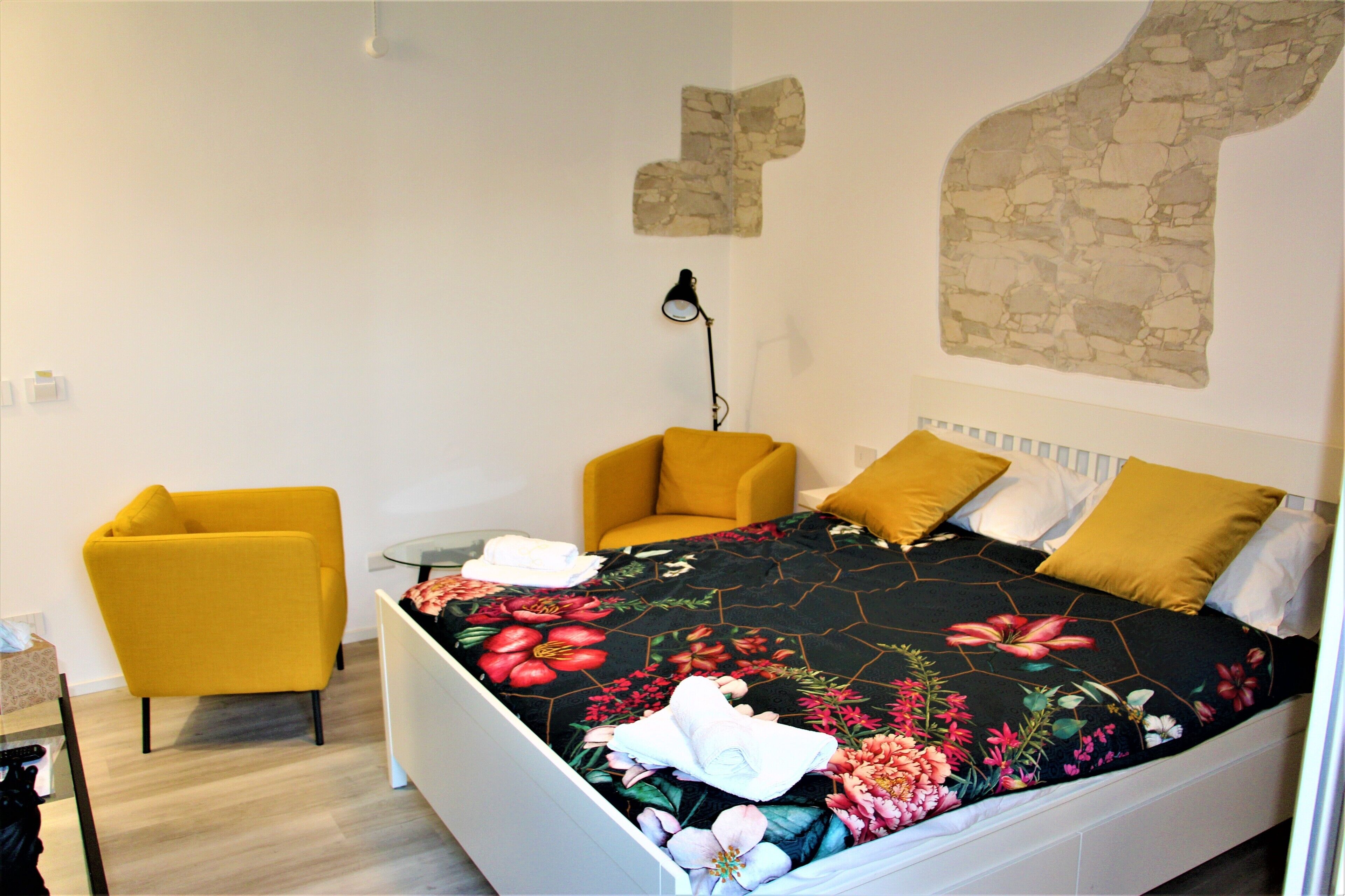 A bedroom with a bed, two yellow chairs, a small table, and a wall-mounted lamp.