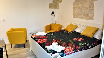 A bedroom with a bed, two yellow chairs, a small table, and a wall-mounted lamp.