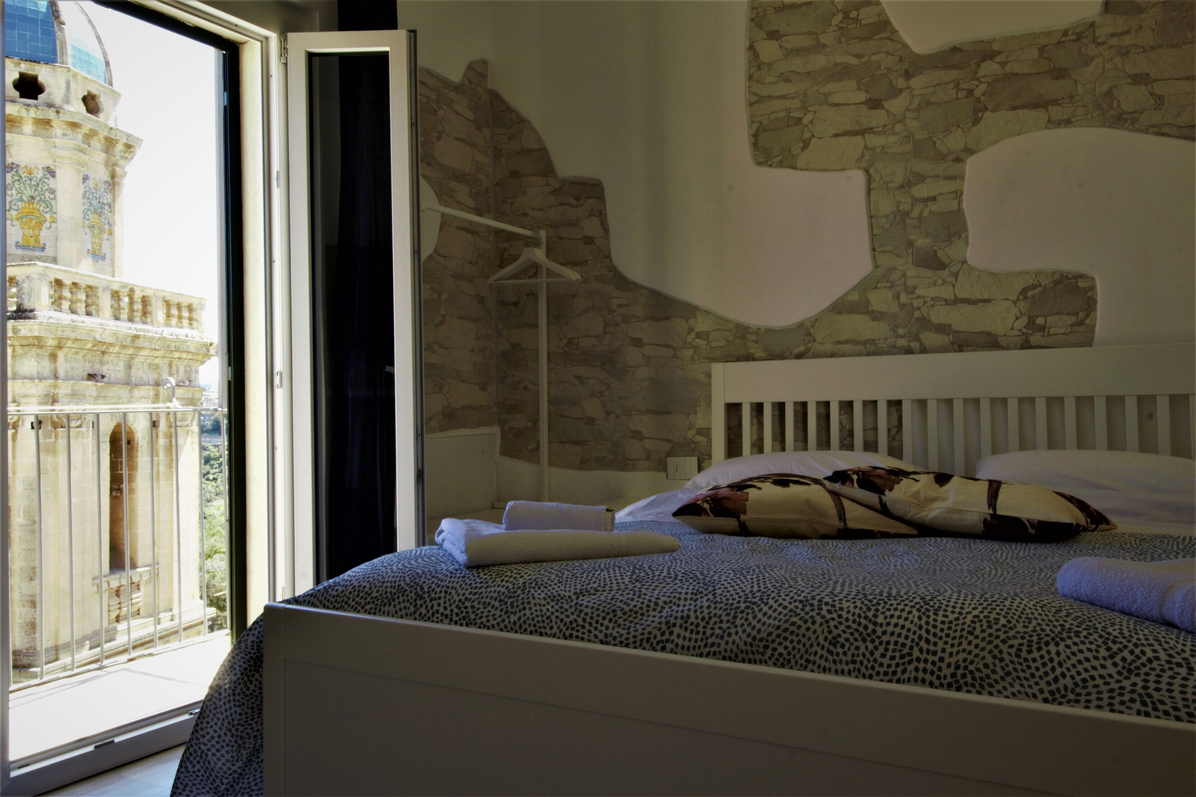 Une chambre à coucher avec un grand lit, une tête de lit blanche et une vue sur un bâtiment historique par une fenêtre ouverte.