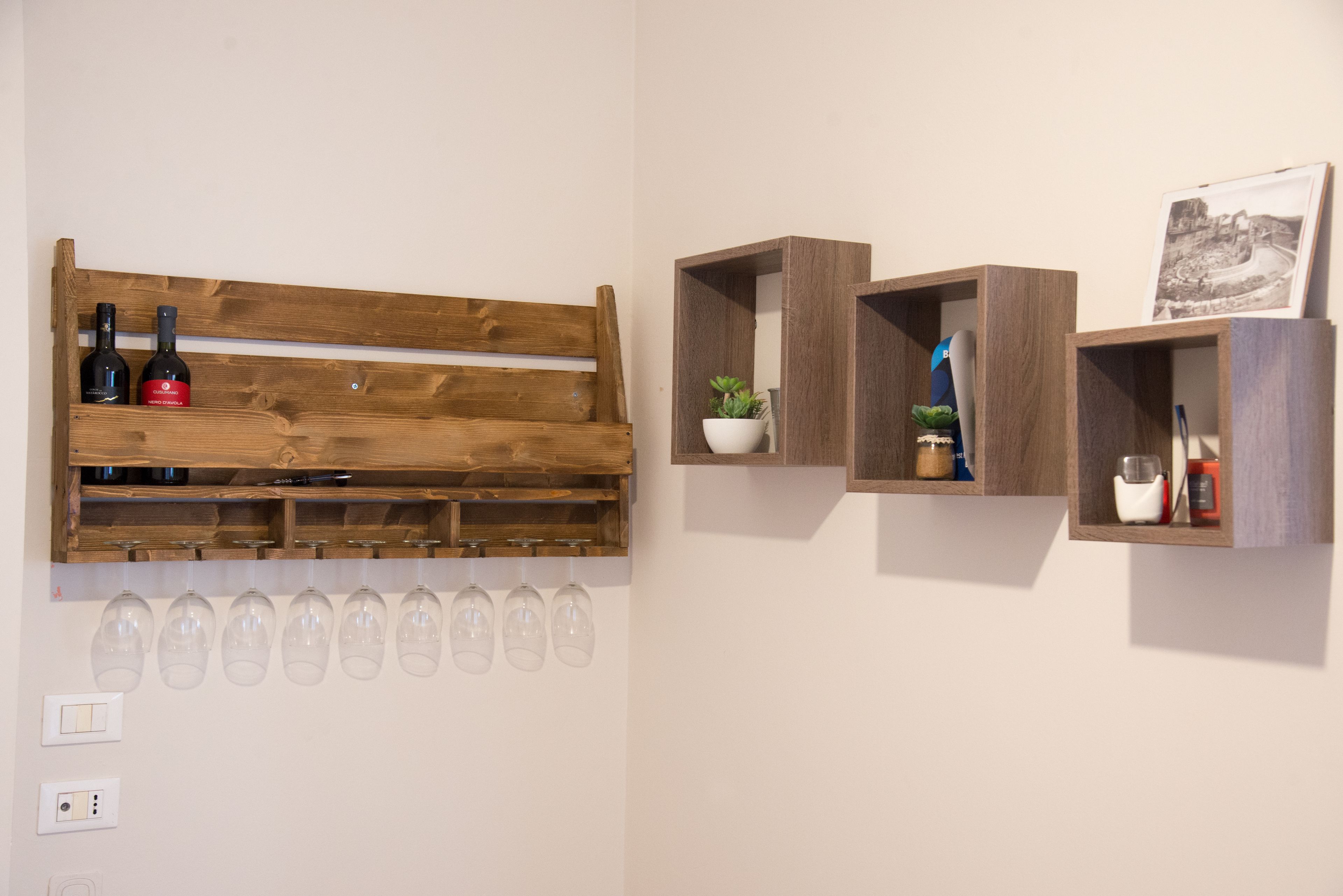 A wall with a wooden bench, wine bottles, glasses, and small wooden shelves with plants and a picture.