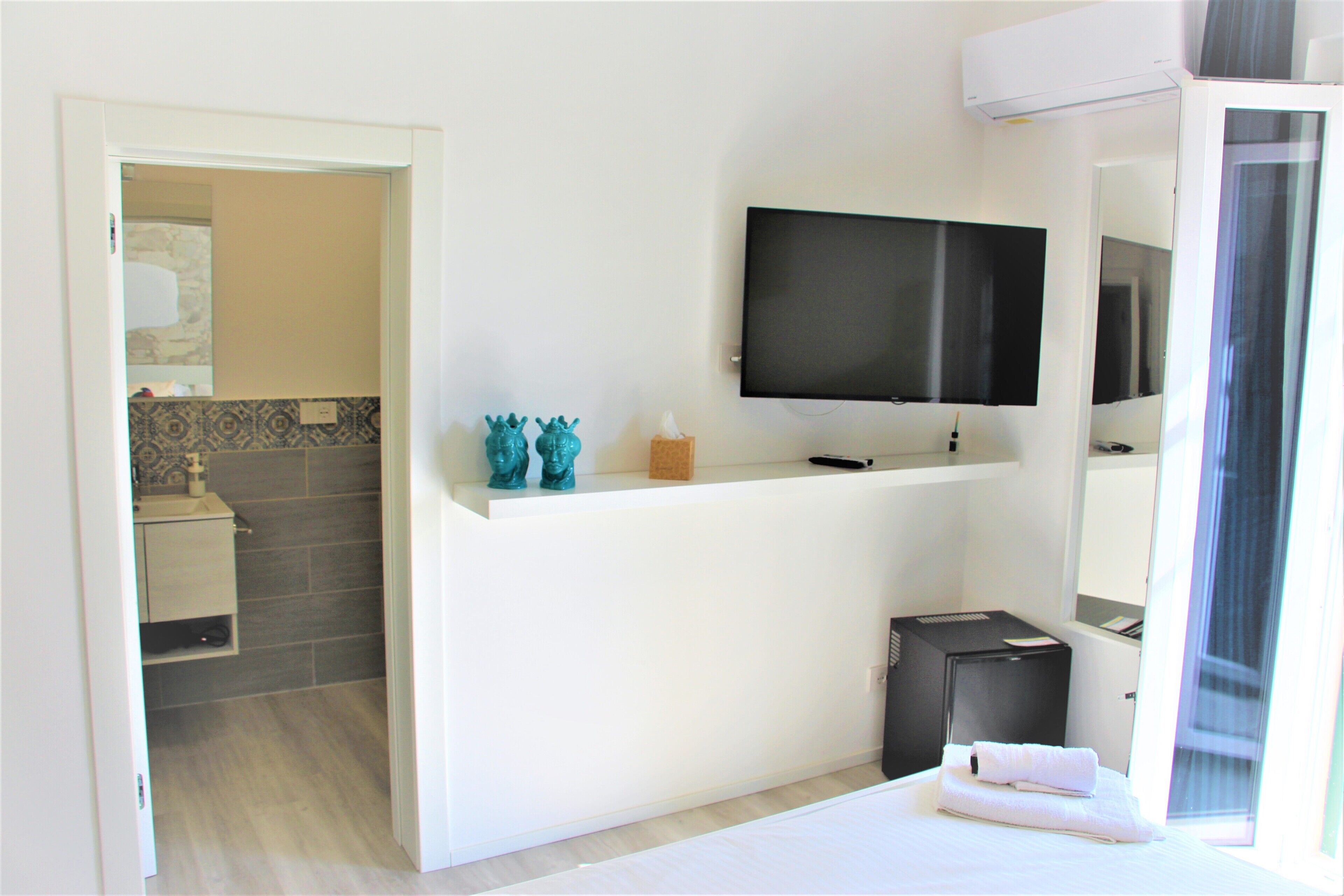 A hotel room with a bed, a TV mounted on the wall, a small desk with a mirror, and a suitcase.