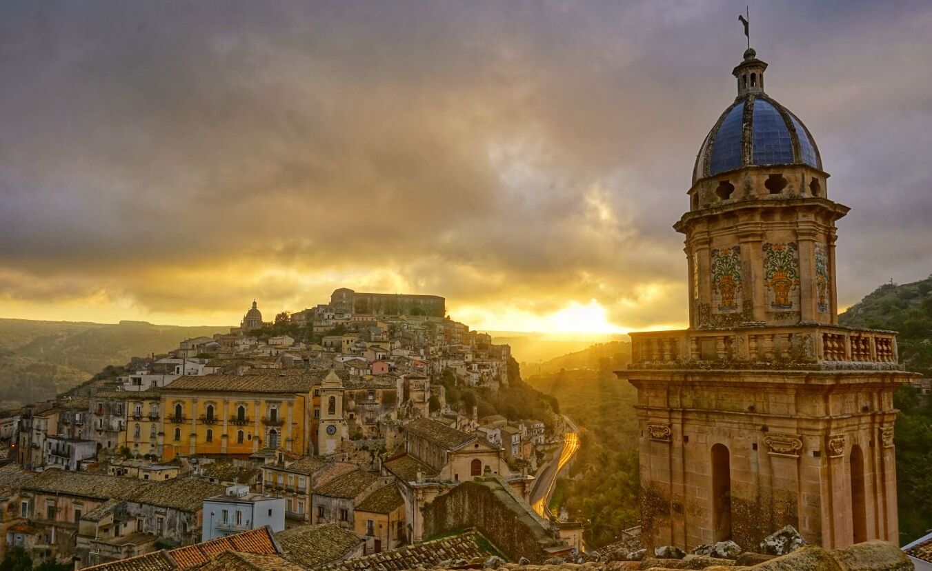 A historic town with a prominent bell tower and a domed building, surrounded by terraced buildings and a mountainous backdrop.
