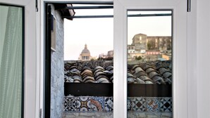 Vista a través de una puerta abierta que muestra un balcón con baldosas, un muro de piedra y un edificio con cúpula al fondo.