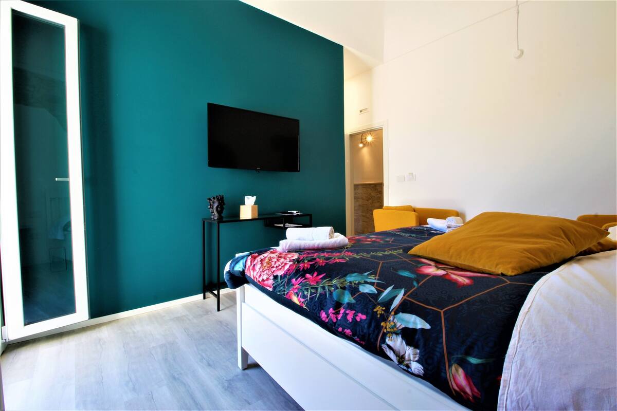Een slaapkamer met een bed, een aan de muur bevestigde televisie, een deur en een klein tafeltje met een lamp.