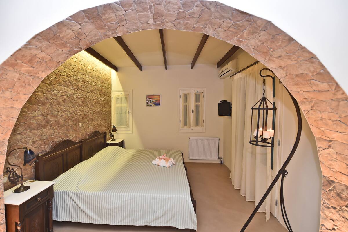 Ein Schlafzimmer mit einem steinernen Bogen, einem Bett mit gestreifter Bettdecke, einem Nachttisch mit Lampe und einer Pendelleuchte.