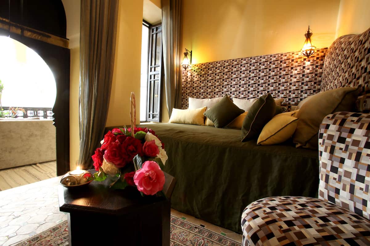 Une chambre à coucher avec une tête de lit à motifs, un lit agrémenté de plusieurs coussins, une table de chevet sur laquelle est posé un vase de fleurs, et une vue à travers une fenêtre en arc de cercle.