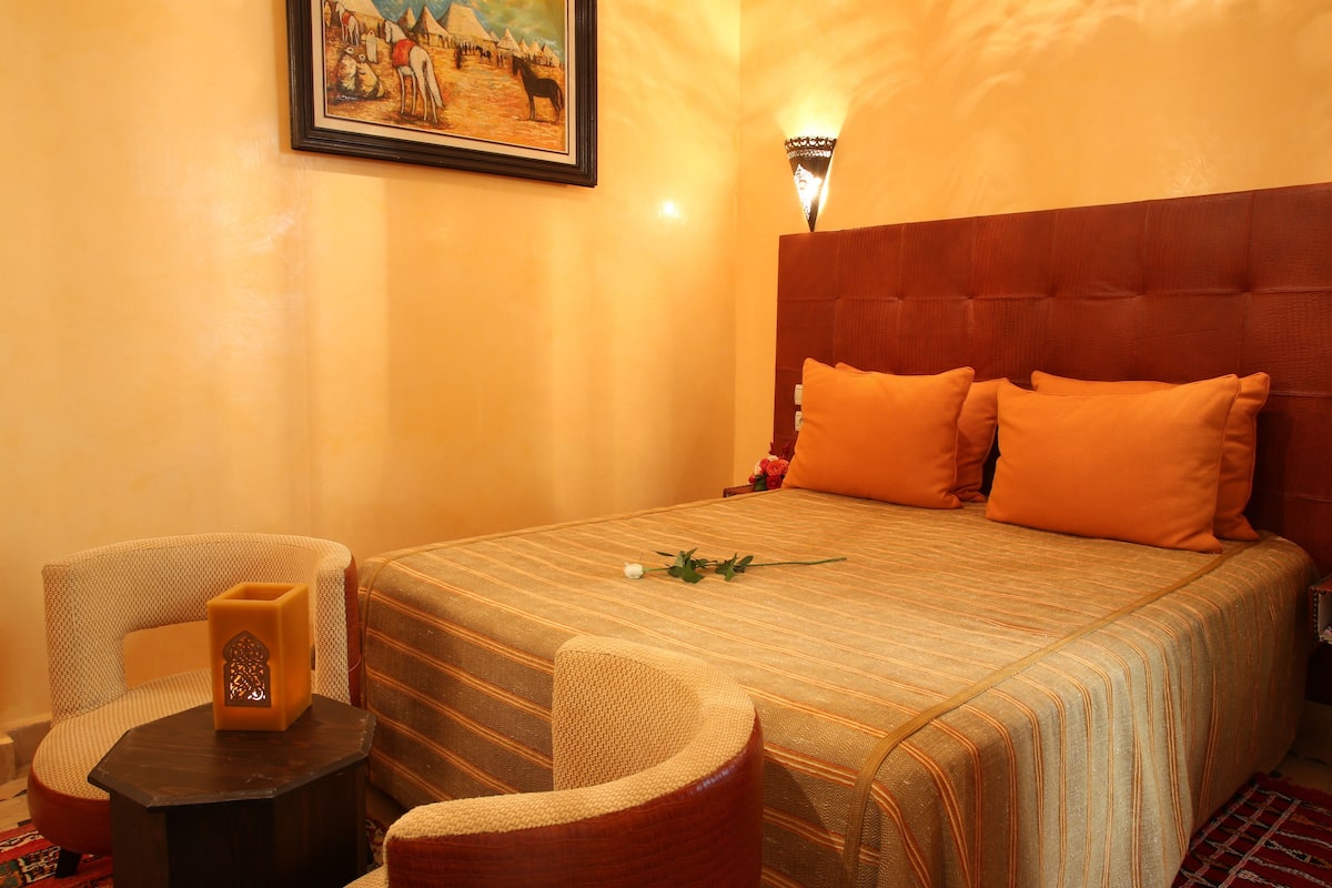 Une chambre d’hôtel avec un grand lit, deux chaises, une petite table, une lampe fixée au mur et un tableau.
