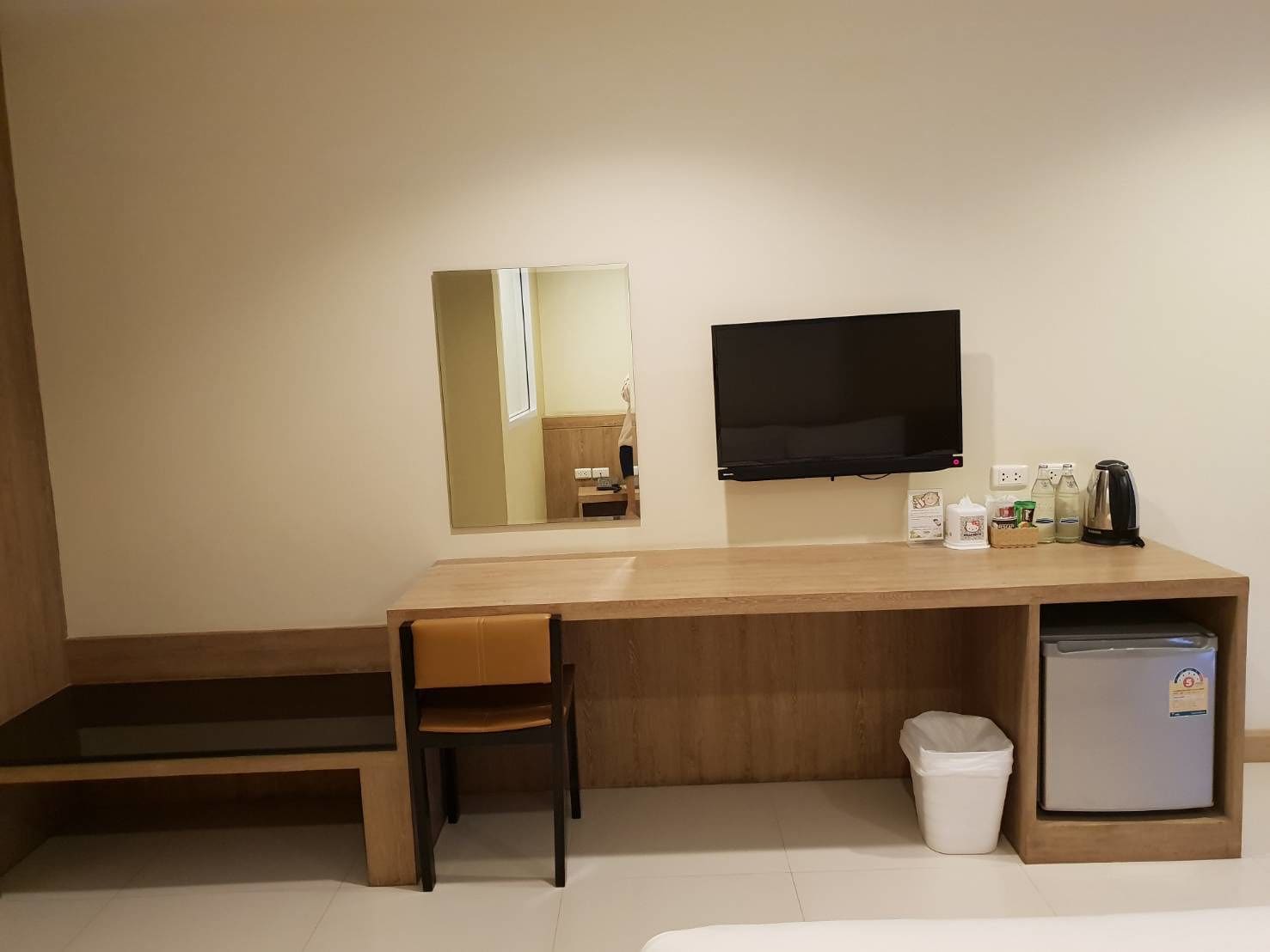 A compact hotel room with a wooden desk, a single chair, a television mounted on the wall, a small refrigerator, and a trash bin.