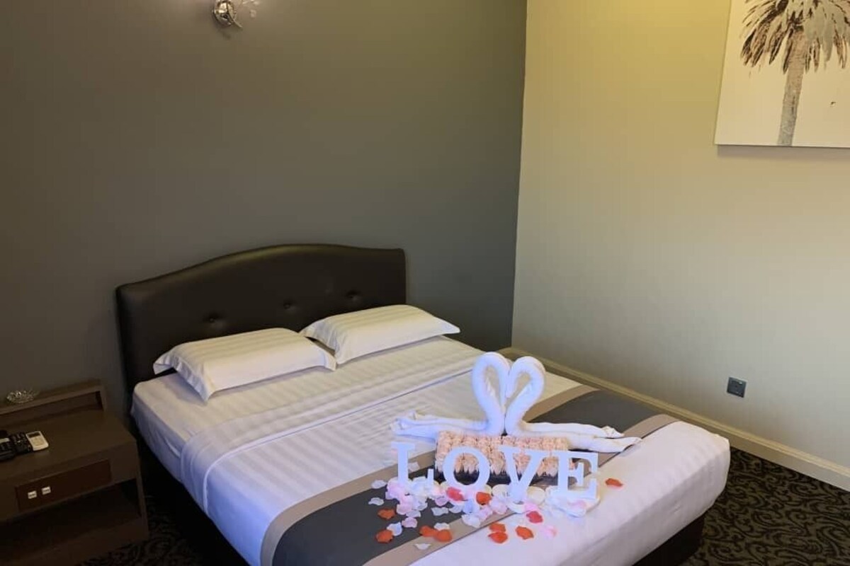 Ein ordentlich bezogenes Bett mit einem herzförmigen Handtuch und der Aufschrift 'LOVE', ein Nachttisch und eine Wandleuchte.