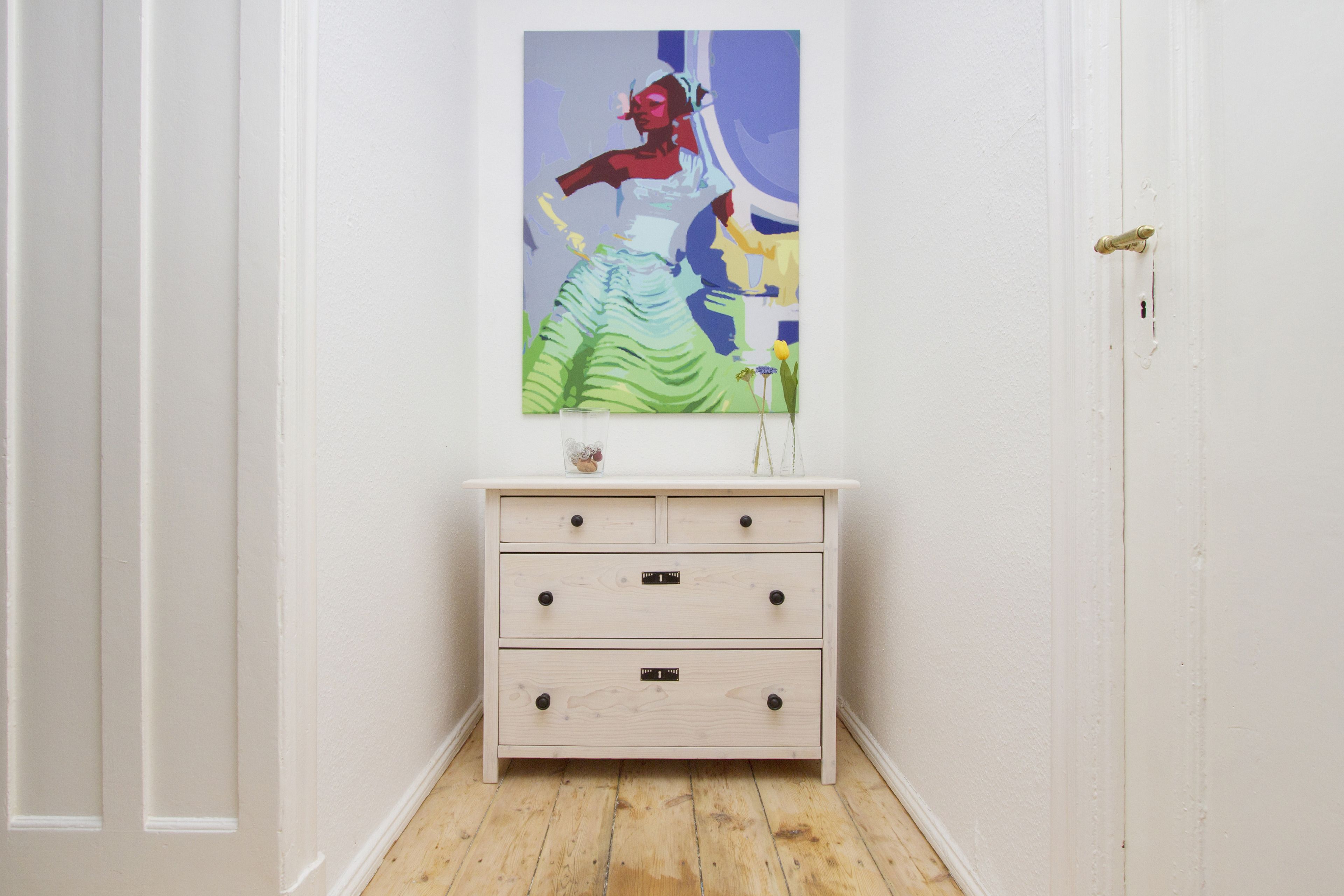 Een gang met een witte deur, een witte dressoir met drie lades, en een kleurrijk schilderij erboven.