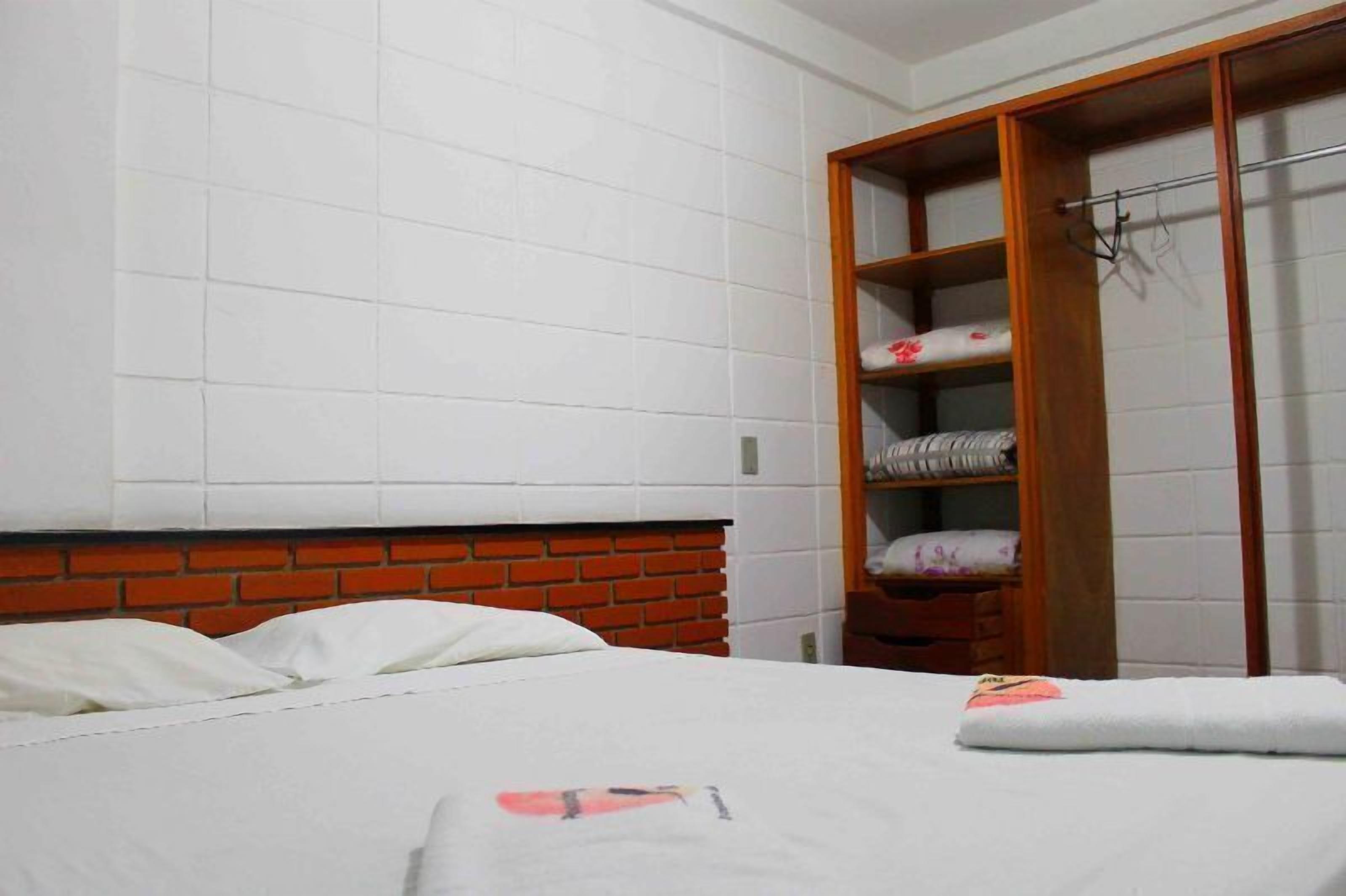 Quarto de hotel com cabeceira de tijolo, paredes com azulejos brancos, guarda-roupa de madeira e roupa de cama bem arrumada.