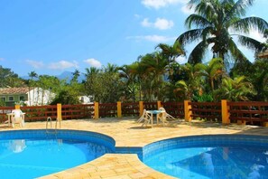 A pool area with two swimming pools, a wooden fence, and palm trees. - Pousada Flat Solar da Tabatinga (Ubatuba)