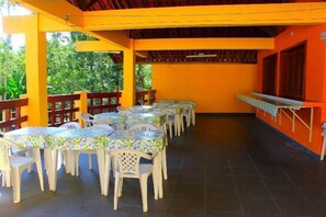 An outdoor dining area with multiple tables and chairs, surrounded by a yellow wall and wooden beams. - Pousada Flat Solar da Tabatinga (Ubatuba)