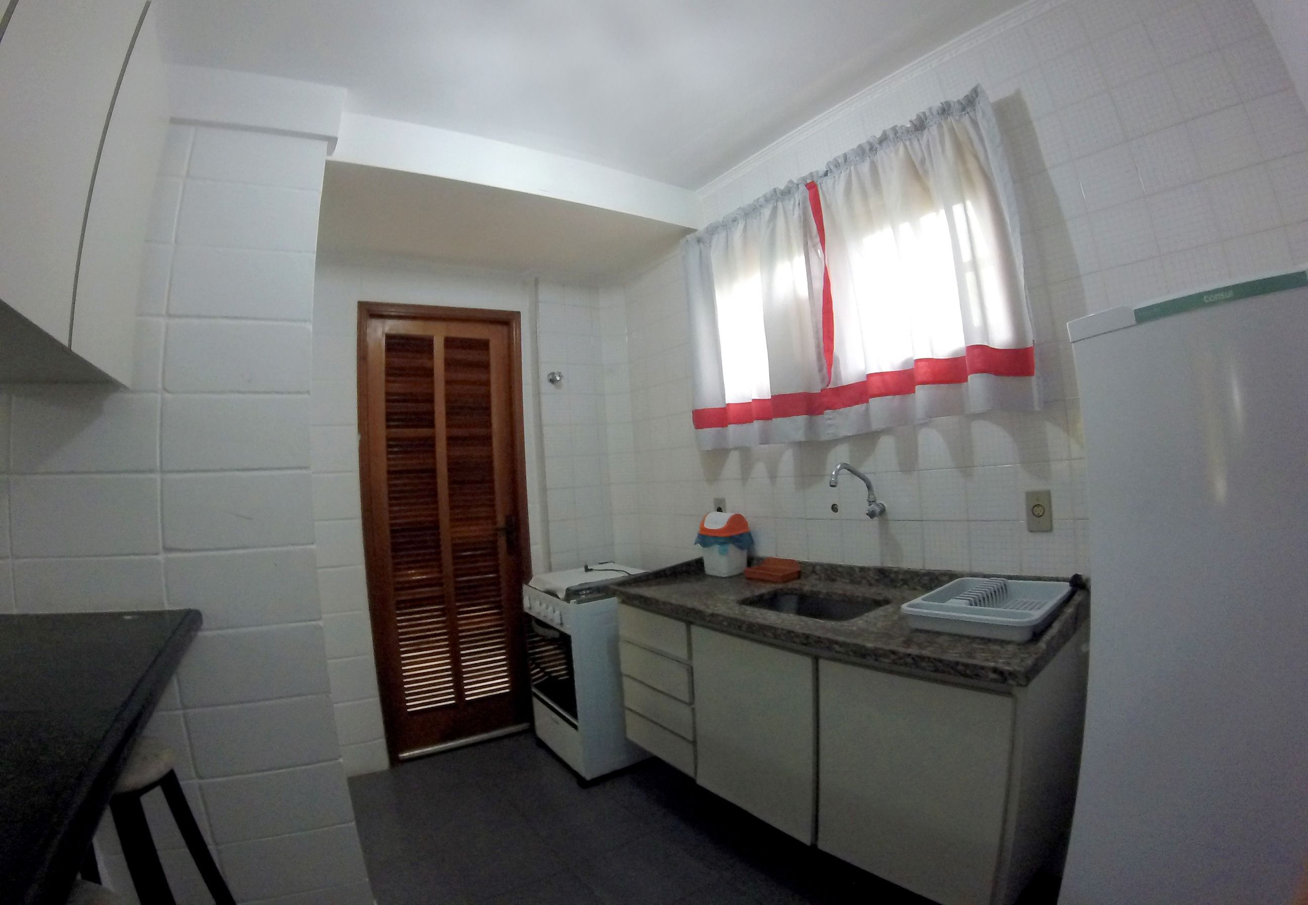 A compact kitchen with white tiled walls, a wooden door, a window with red curtains, a sink, and a countertop.