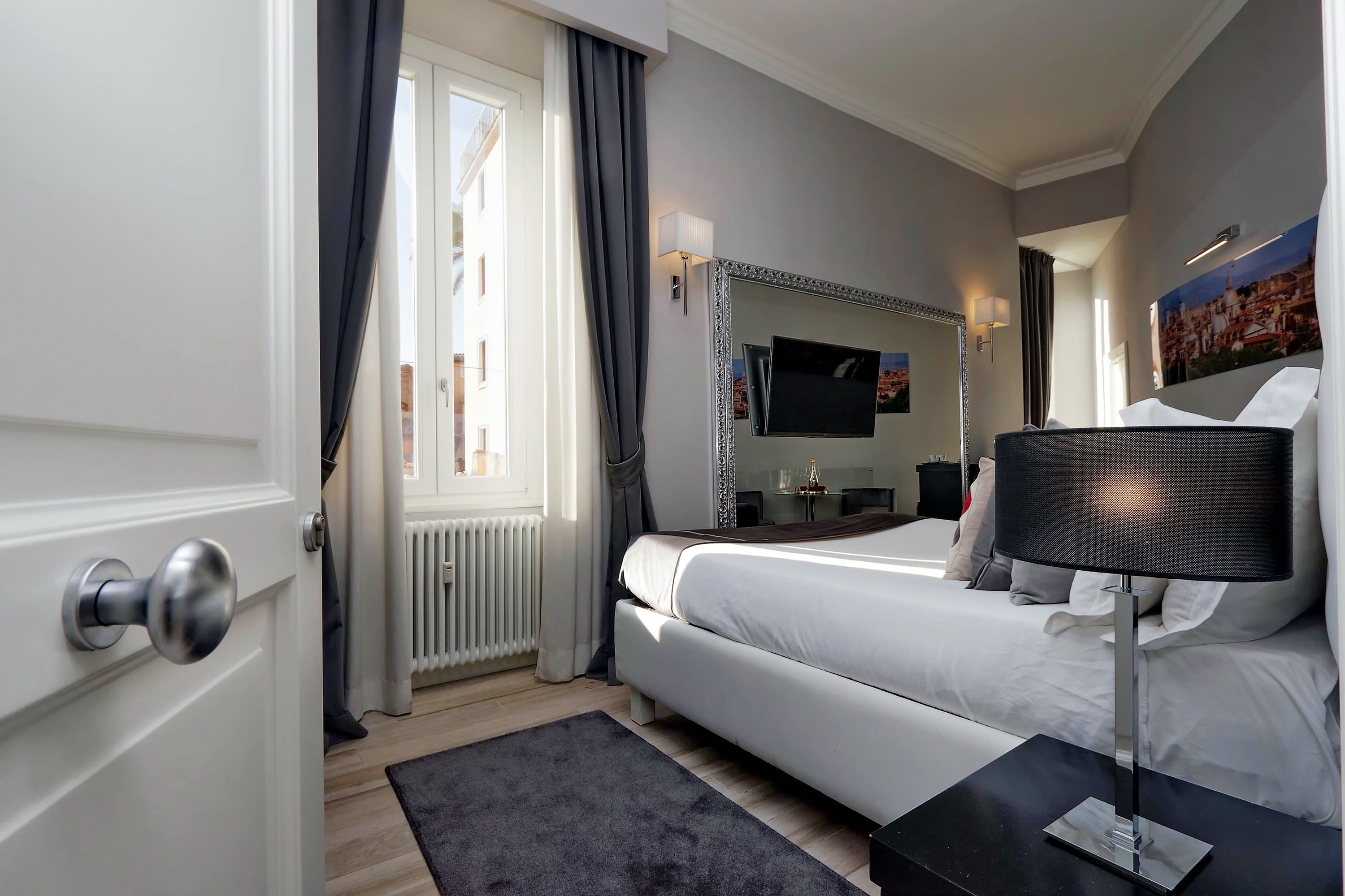 A modern hotel room with a large bed, a bedside table with a lamp, a TV, and a window with curtains.