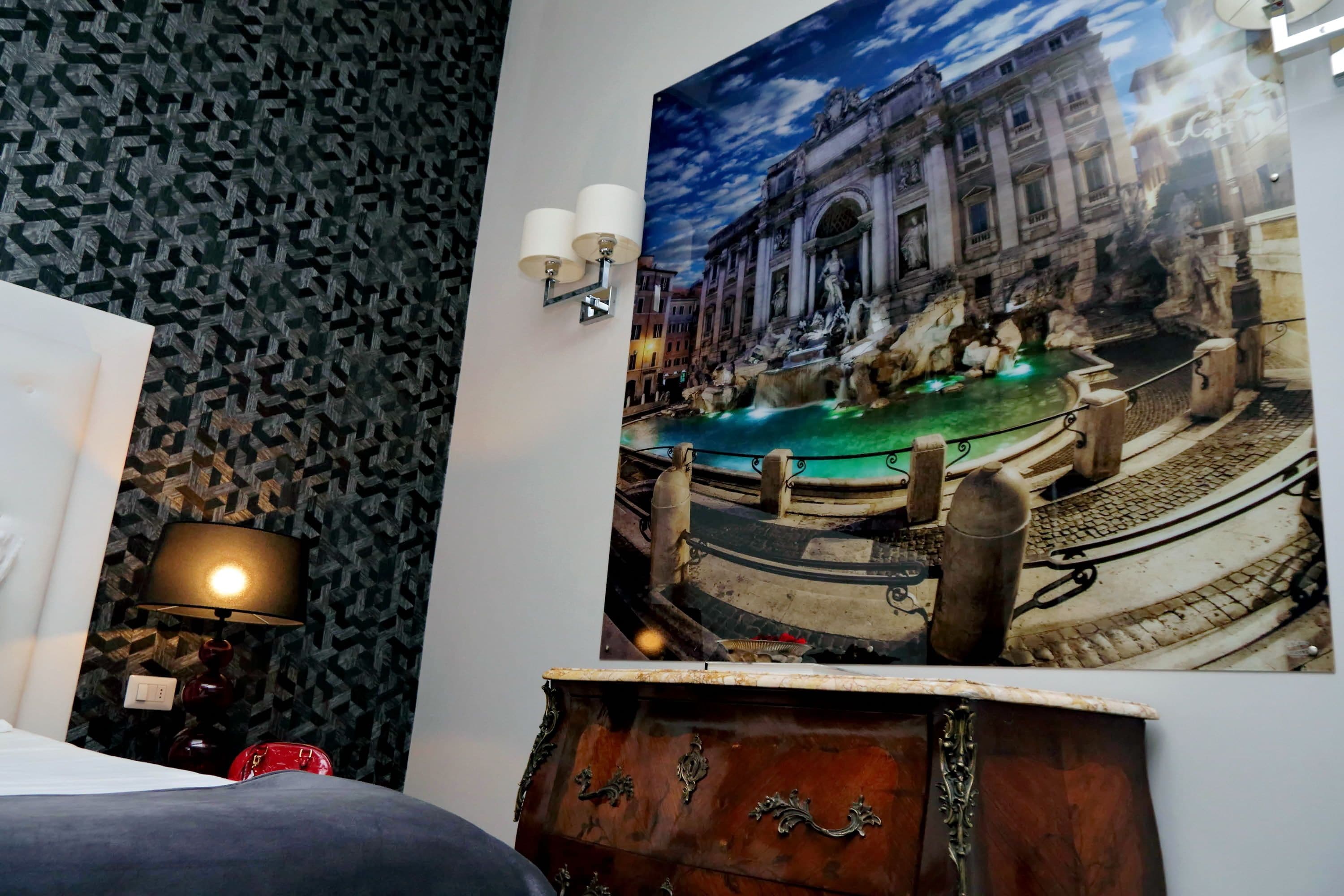 Une chambre d’hôtel avec un papier peint à motifs, un lit, une lampe, un luminaire fixé au mur et une grande fresque représentant une fontaine historique.