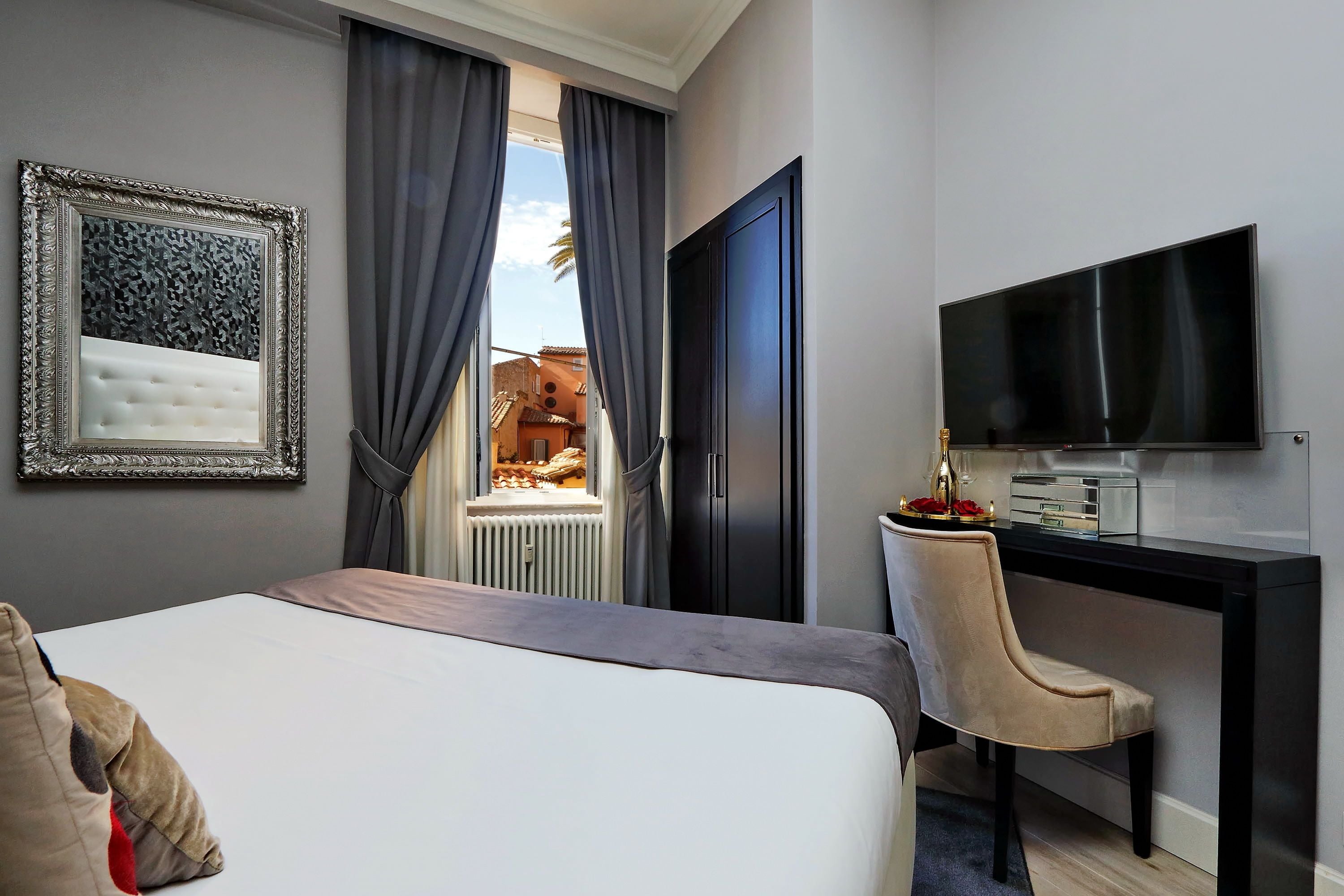 A hotel room with a bed, a desk with a flat-screen TV, a chair, and a view of a balcony with a palm tree.