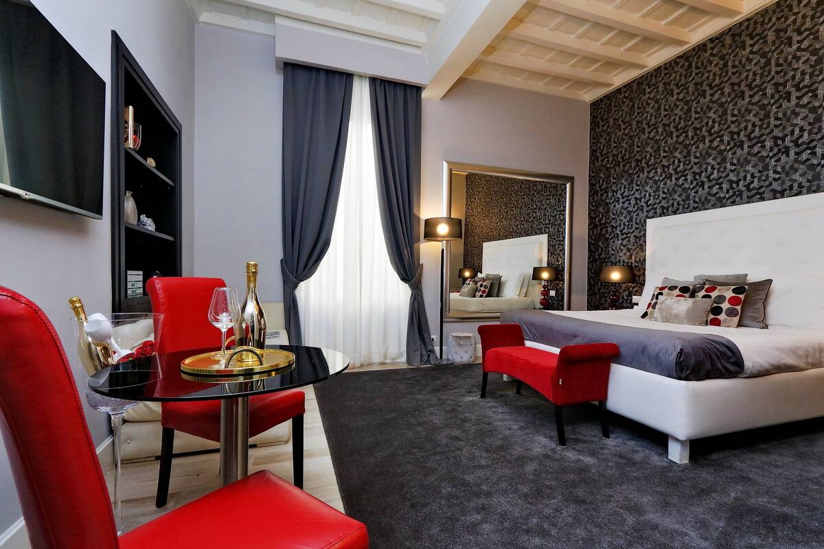A modern hotel room with a large bed, a dining area with a glass table and red chairs, a bookshelf, and a large mirror.
