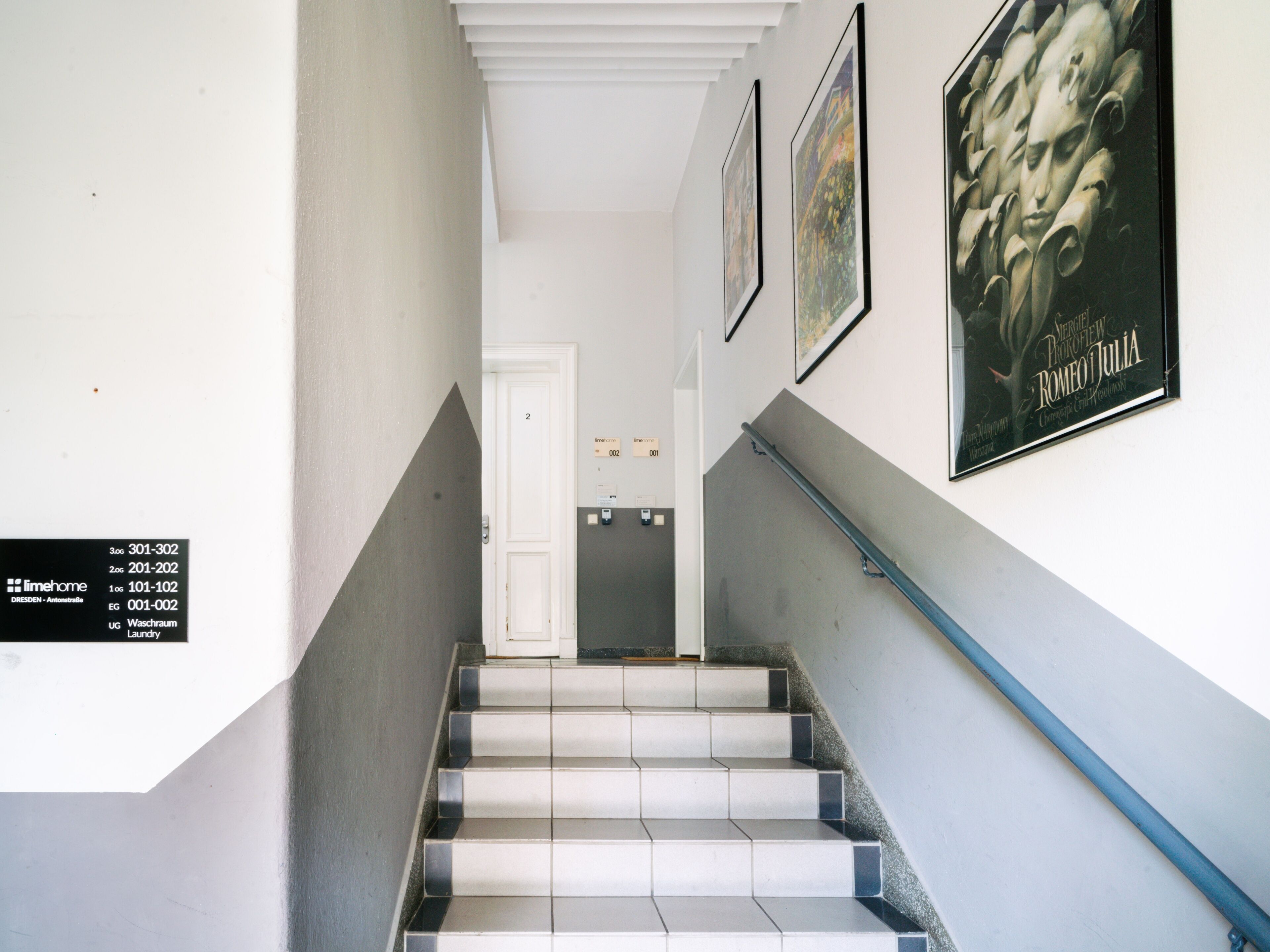 A staircase with white and grey tiles, a blue handrail, and a door at the top. There are framed pictures on the walls and a sign with room numbers.
