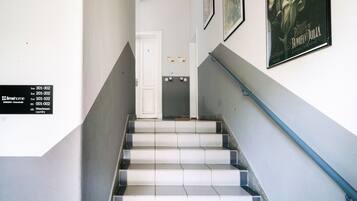 A staircase with white and grey tiles, a blue handrail, and a door at the top. There are framed pictures on the walls and a sign with room numbers.