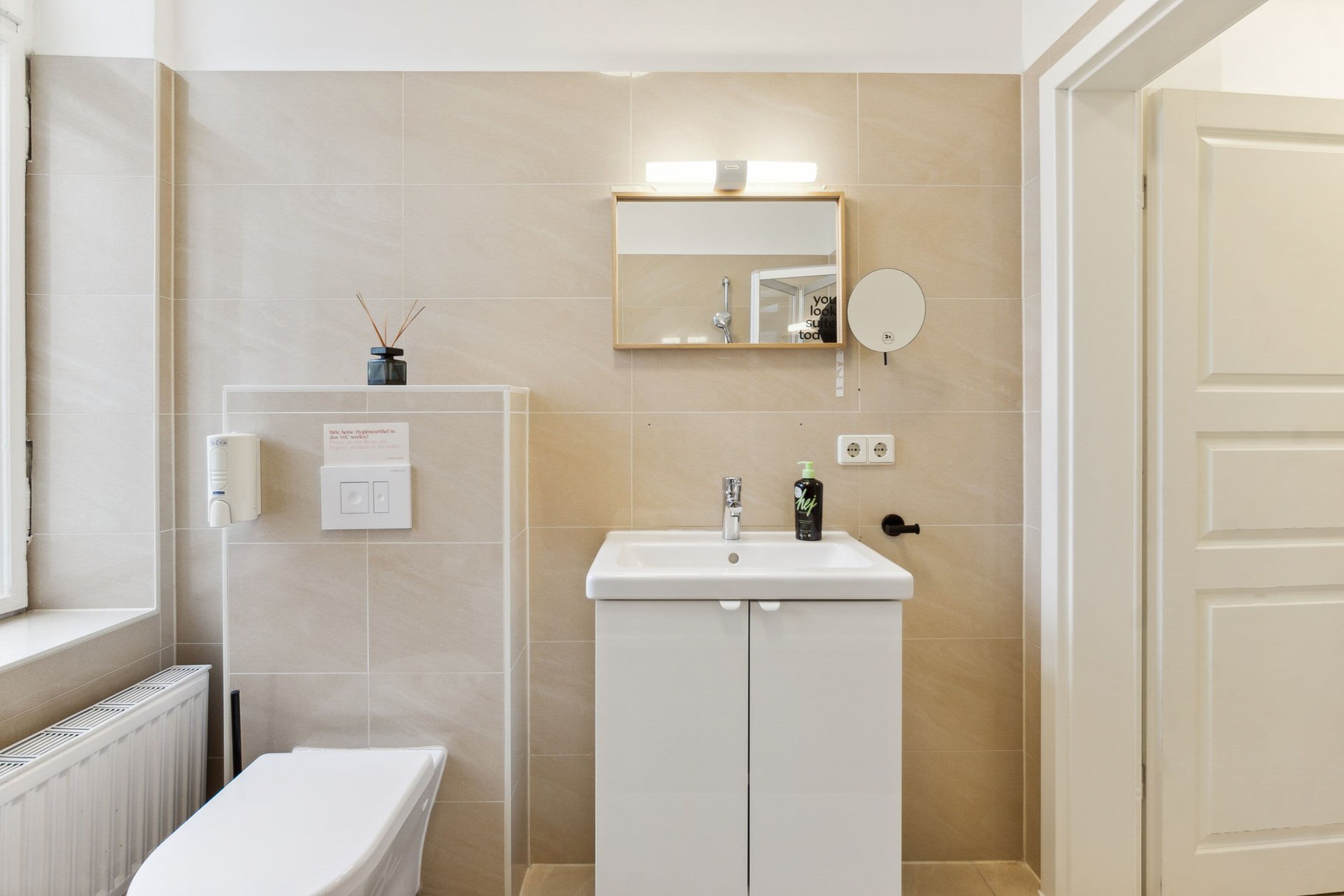 Un baño con lavamanos, espejo y retrete blancos. Se observa una ventana a la izquierda y una puerta a la derecha.