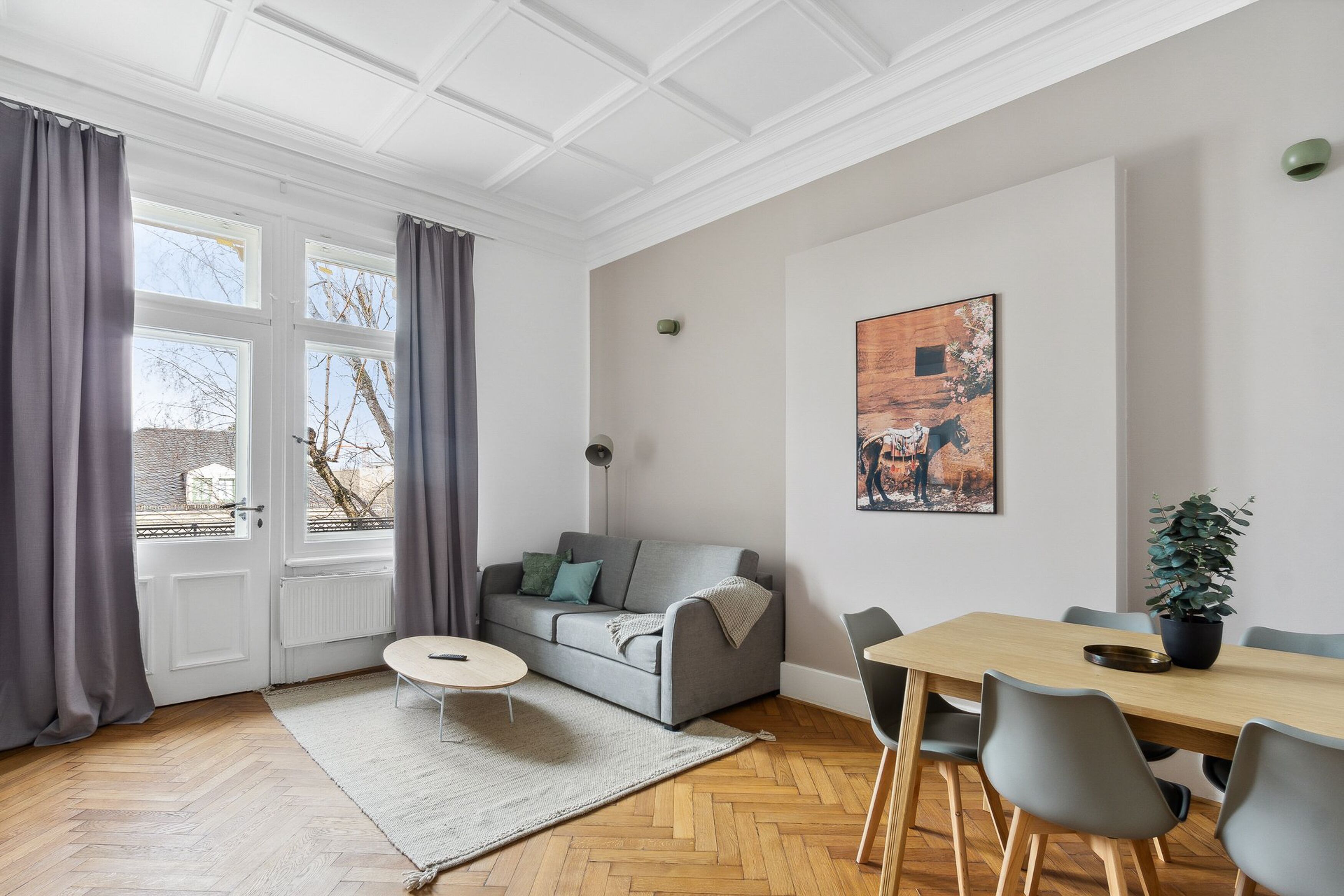 A modern living room with a grey sofa, a round coffee table, a dining table with chairs, and a potted plant.