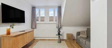 Una sala de estar moderna con televisor de pantalla plana, consola de madera, sofá gris y vista al cielo a través de ventanas.