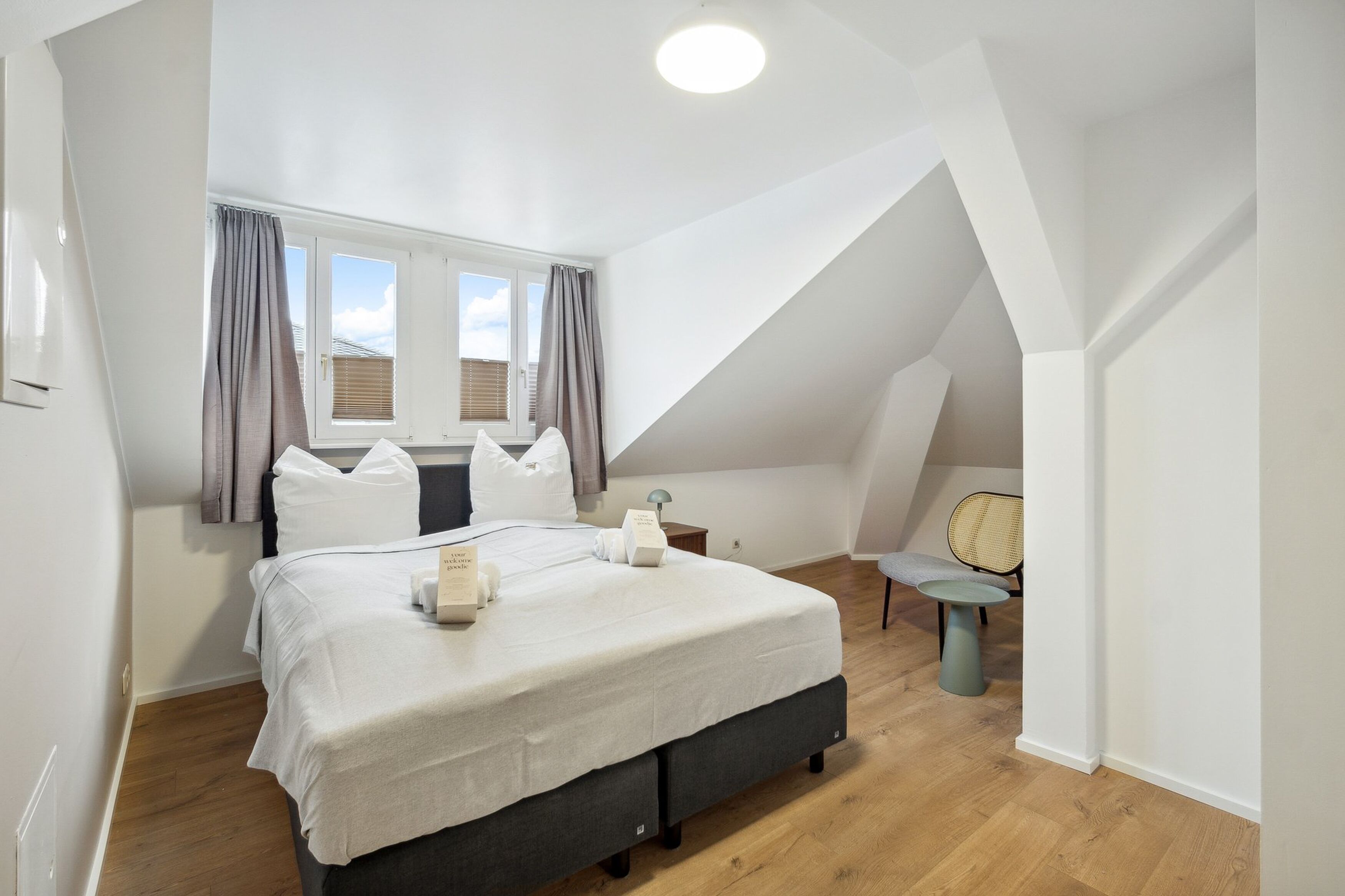A bedroom with a large bed, a window with curtains, a small table, and a chair.