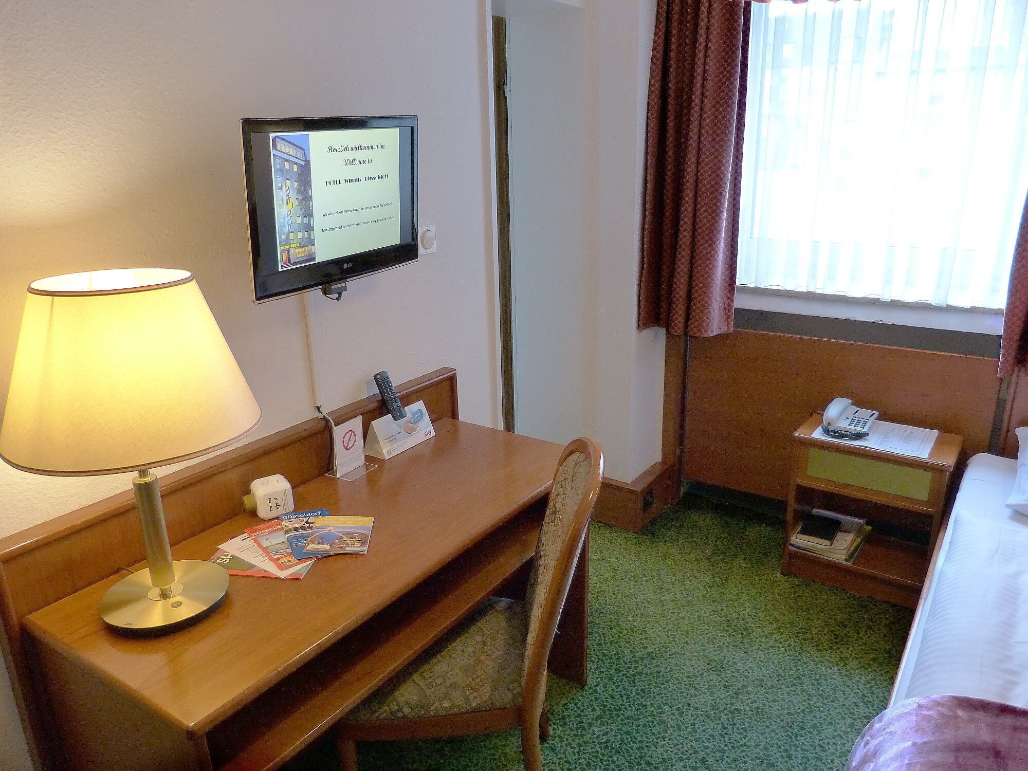 A hotel room with a desk, a lamp, a television displaying a welcome message, a bed, and a window with curtains.