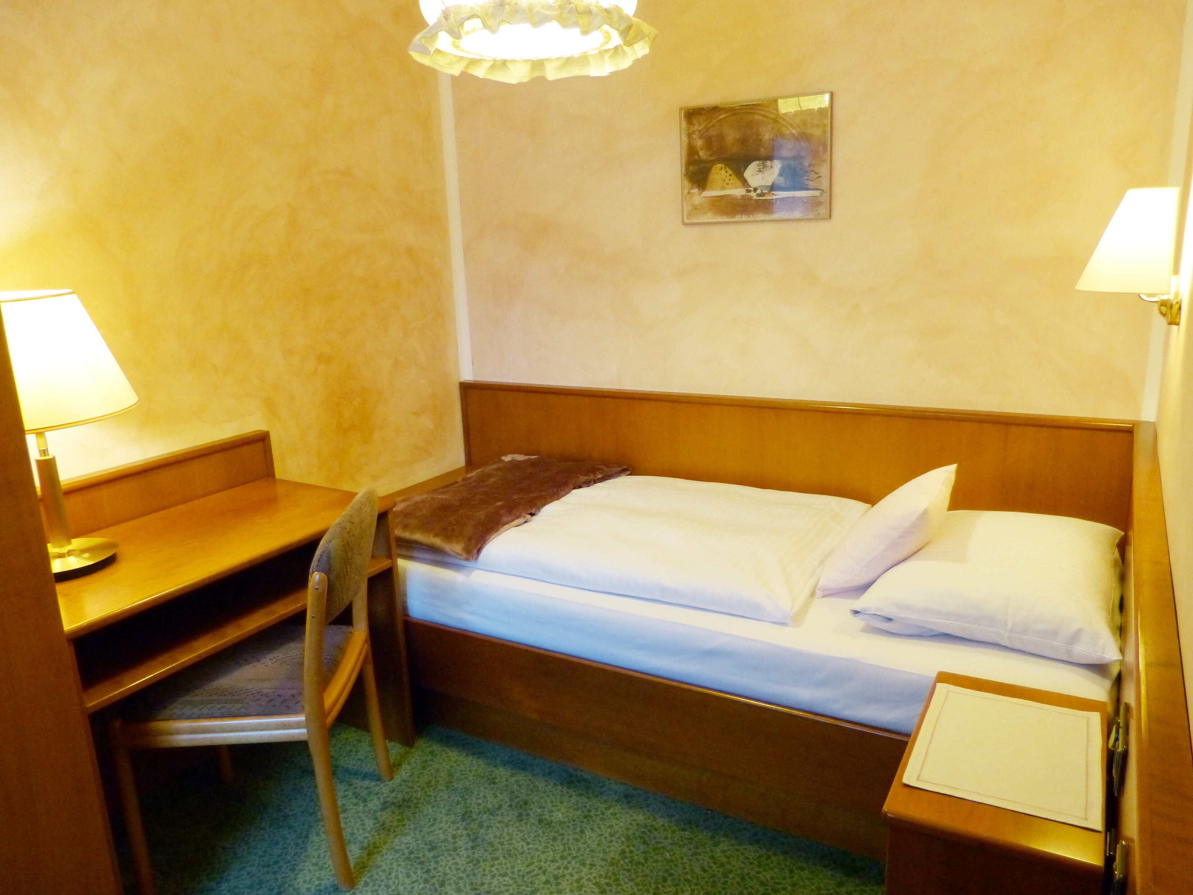 A small hotel room with a single bed, a desk, and a chair. There is a painting on the wall and a lamp on the desk.