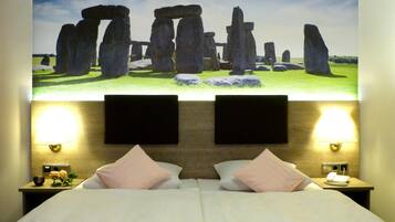 A hotel room with a bed, two bedside lamps, a nightstand with a phone, and a large wall mural of Stonehenge.