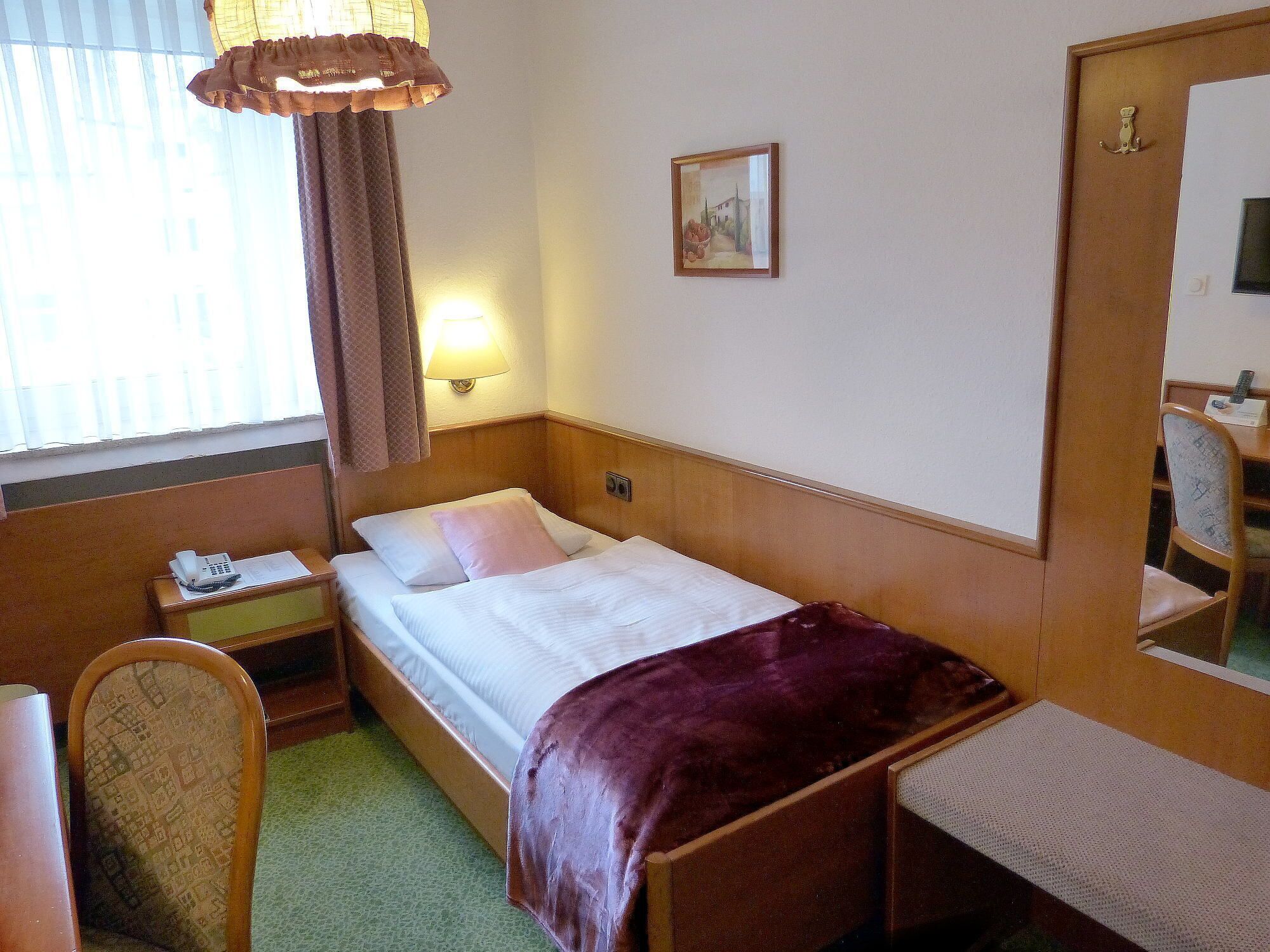 A hotel room with a single bed, a bedside table with a lamp, a chair, a desk with a telephone, and a window with curtains.