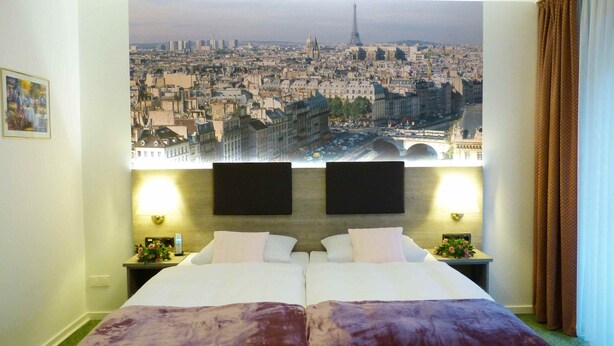 A hotel room with a large bed, two bedside tables, a nightstand, a lamp, and a view of a cityscape. - Hotel Wurms (Düsseldorf)