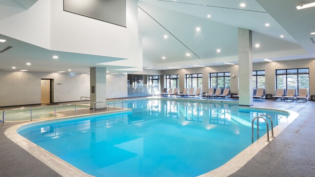 Une piscine intérieure à l’eau d’un bleu cristallin, entourée de chaises longues et dotée de grandes fenêtres.