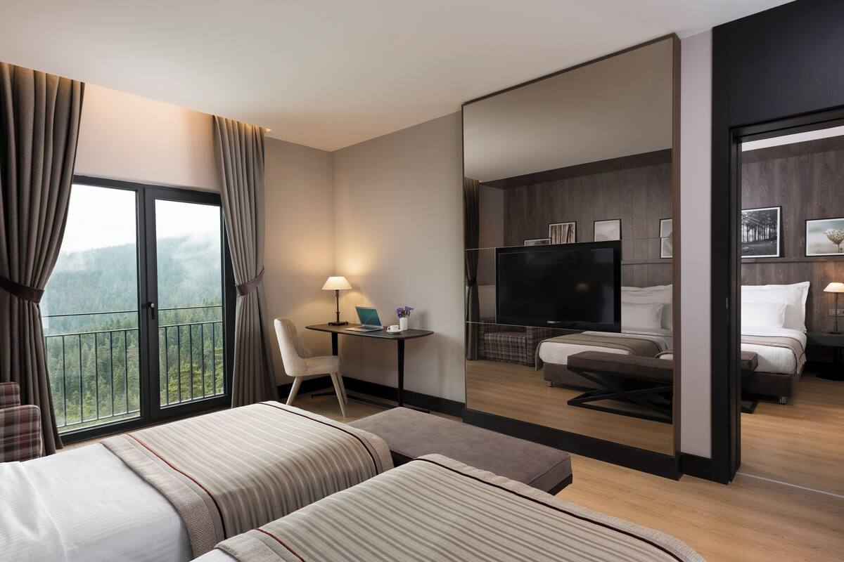 Une chambre d’hôtel avec deux lits, un bureau avec un ordinateur portable, une télévision et un balcon avec des rideaux.