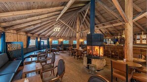 Un intérieur rustique en bois, avec un poêle à bois central, des coins salon et une lumière naturelle provenant des fenêtres.