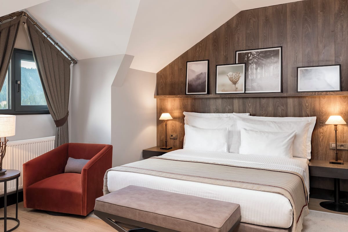 Une chambre à coucher moderne avec un lit, un fauteuil rouge, une table de chevet avec une lampe et une fenêtre avec des rideaux.