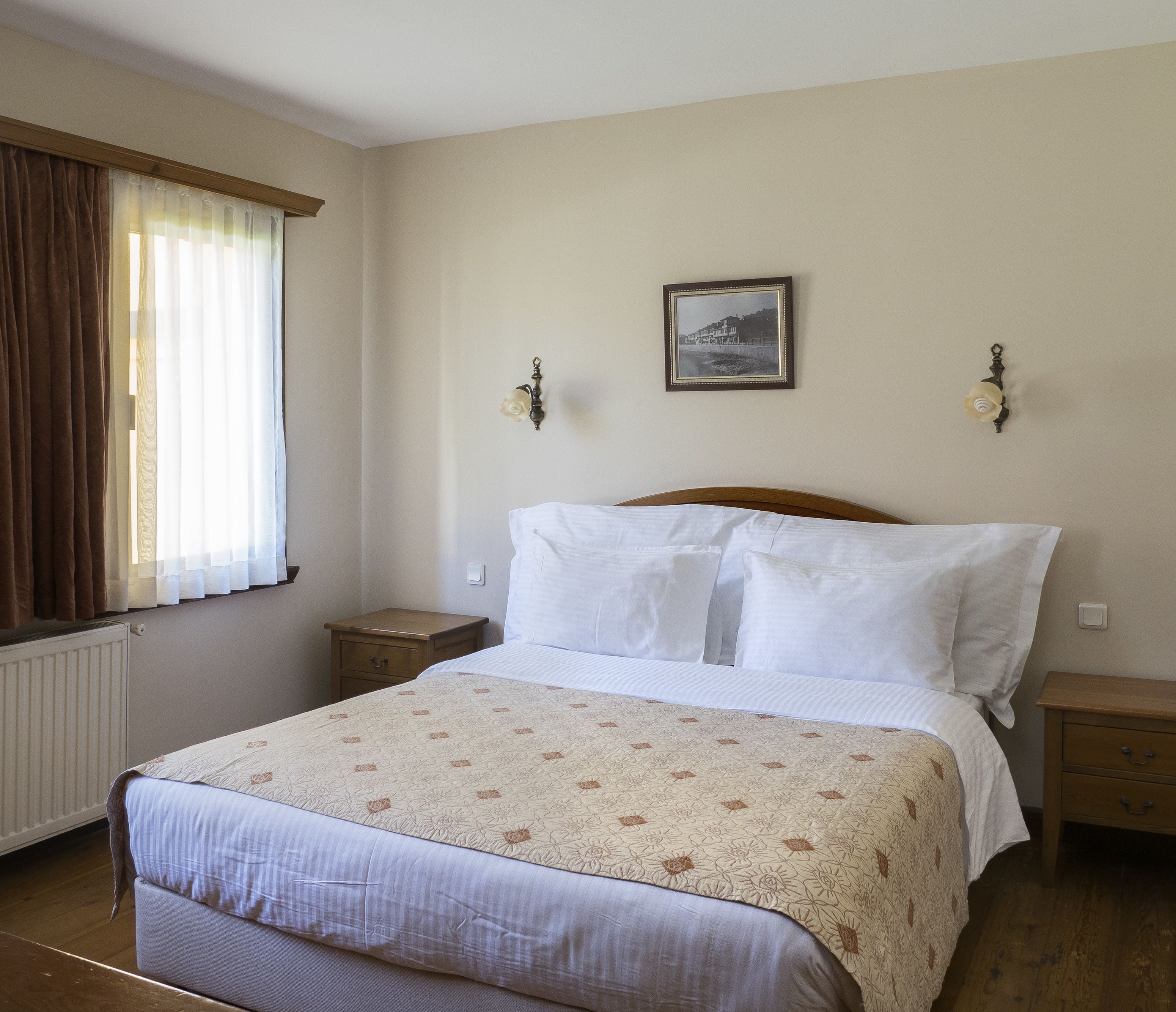 A neatly made bed with a floral-patterned bedspread, two bedside tables, a nightstand, a framed picture on the wall, and a window with curtains.