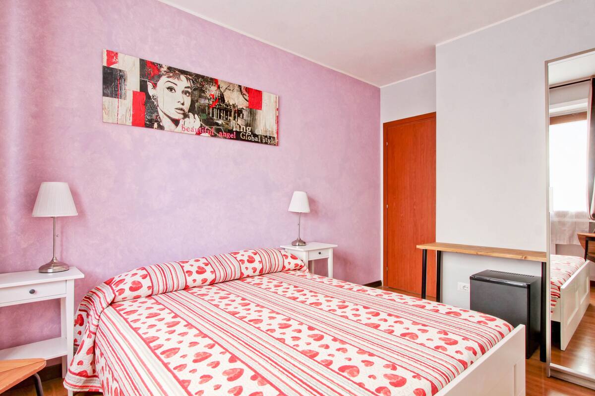 A bedroom with a pink wall, a bed with a red and white patterned bedspread, two bedside tables with lamps, a small desk, a mirror, and a wardrobe.