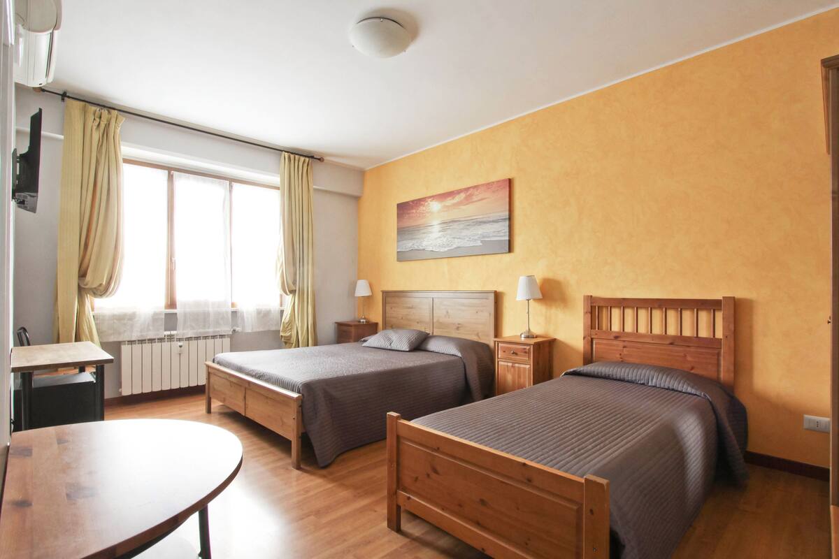 A hotel room with two beds, a wooden table, a window with curtains, a painting on the wall, and a ceiling light.