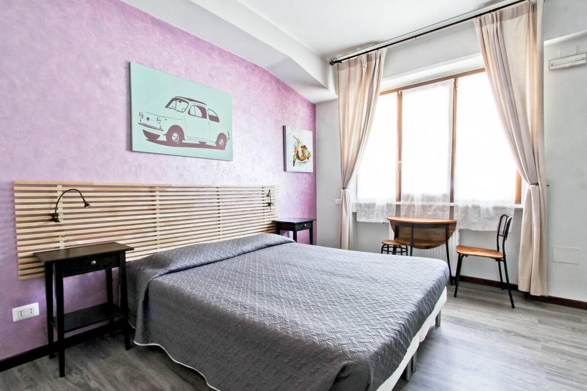 A bedroom with a bed, a desk, a chair, a window with curtains, and artwork on the wall.