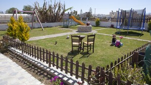 A fenced backyard with a grassy lawn, a play area including a slide and swing set, and a small table and chairs. - Villa Bel Passo (Kos)