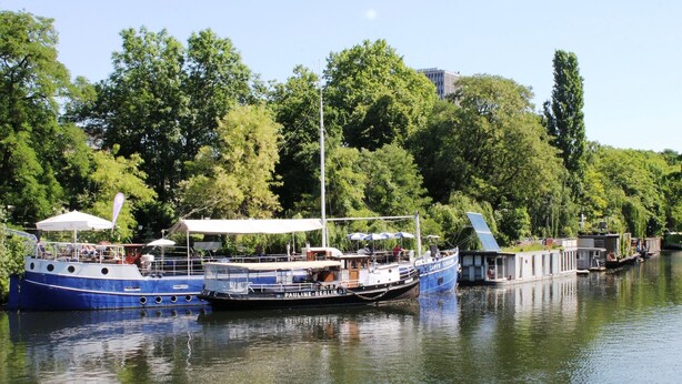 A row of boats docked along a river, with a canopy-covered area and a building with a sloped roof. - Lovely Flats Englische Straße (Berlin)