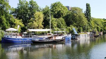 A row of boats docked along a river, with a canopy-covered area and a building with a sloped roof.