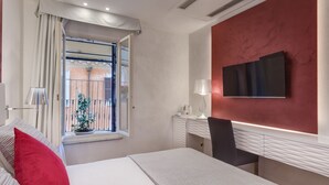 A hotel room with a bed, a desk, a chair, a TV, a window with curtains, and a potted plant. - Shali Luxury Suite (Rome)