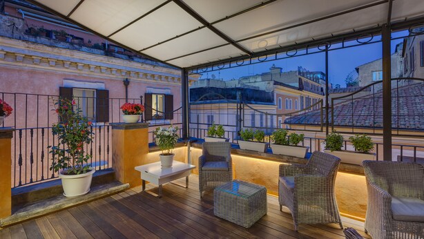 A covered outdoor seating area with wicker furniture, potted plants, and a view of city buildings. - Shali Luxury Suite (Rome)