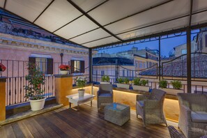 A covered outdoor seating area with wicker furniture, potted plants, and a view of city buildings. - Shali Luxury Suite (Rome)