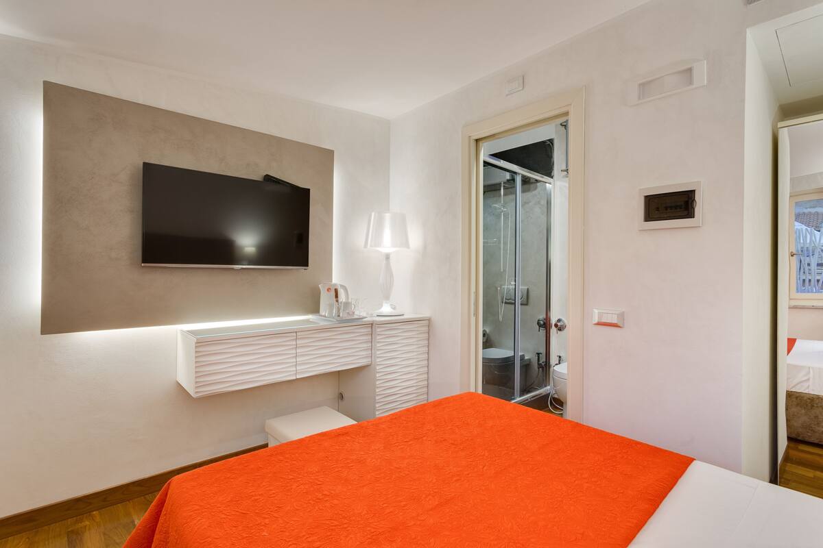 A hotel room with a bed, a TV mounted on the wall, a bathroom with a glass shower enclosure, and a white bedside table with a lamp.